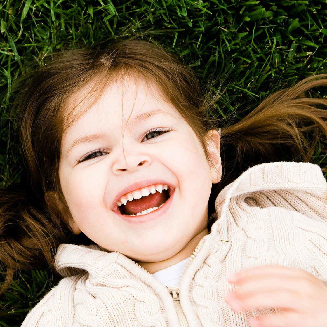 A little girl in a cream cable-knit cardigan lying on the grass, laughing; every strand of hair, blade of grass and stitch of her cardigan is sharp.