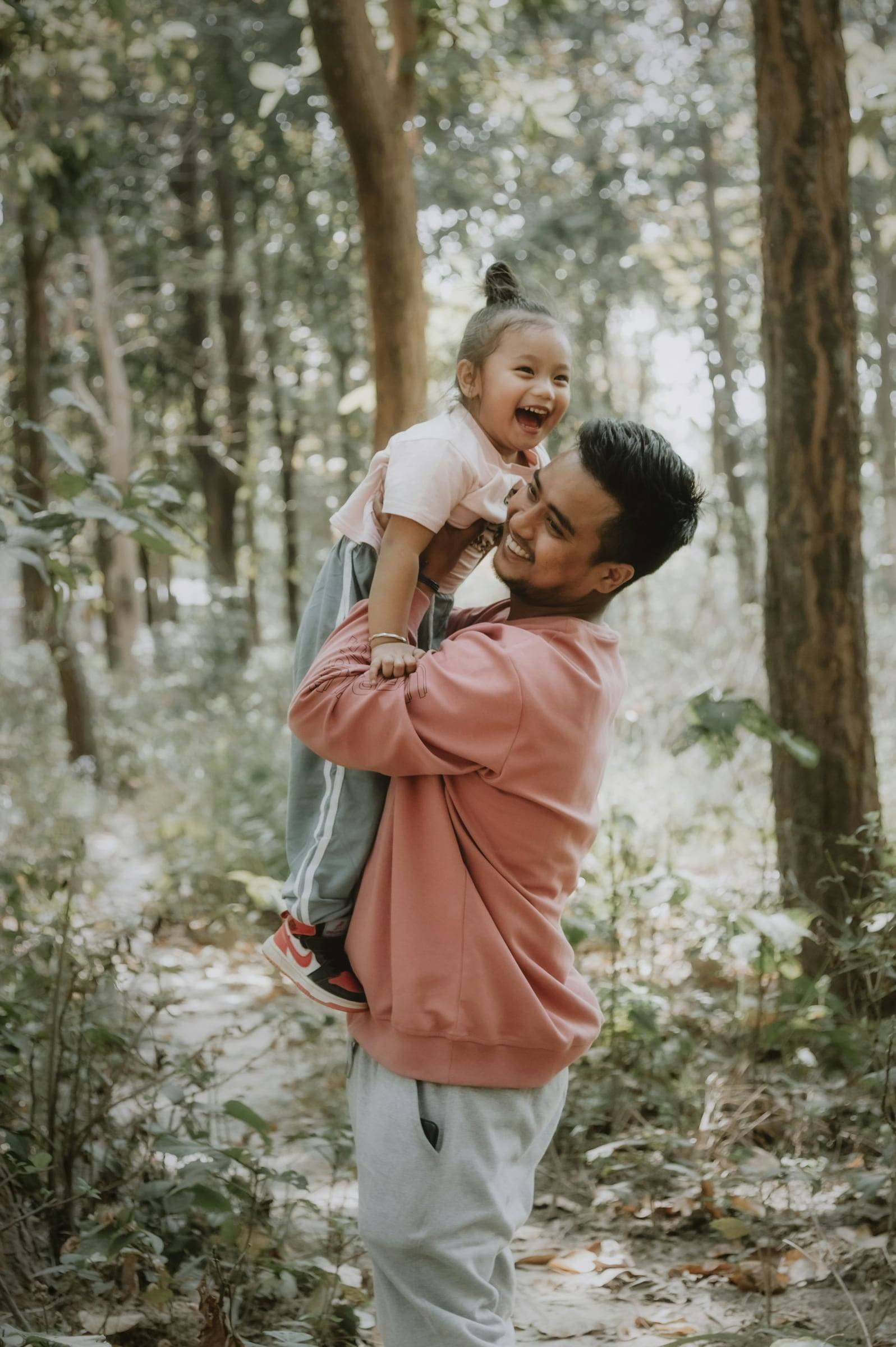 A father lifting his laughing little daughter into the air in a forest.