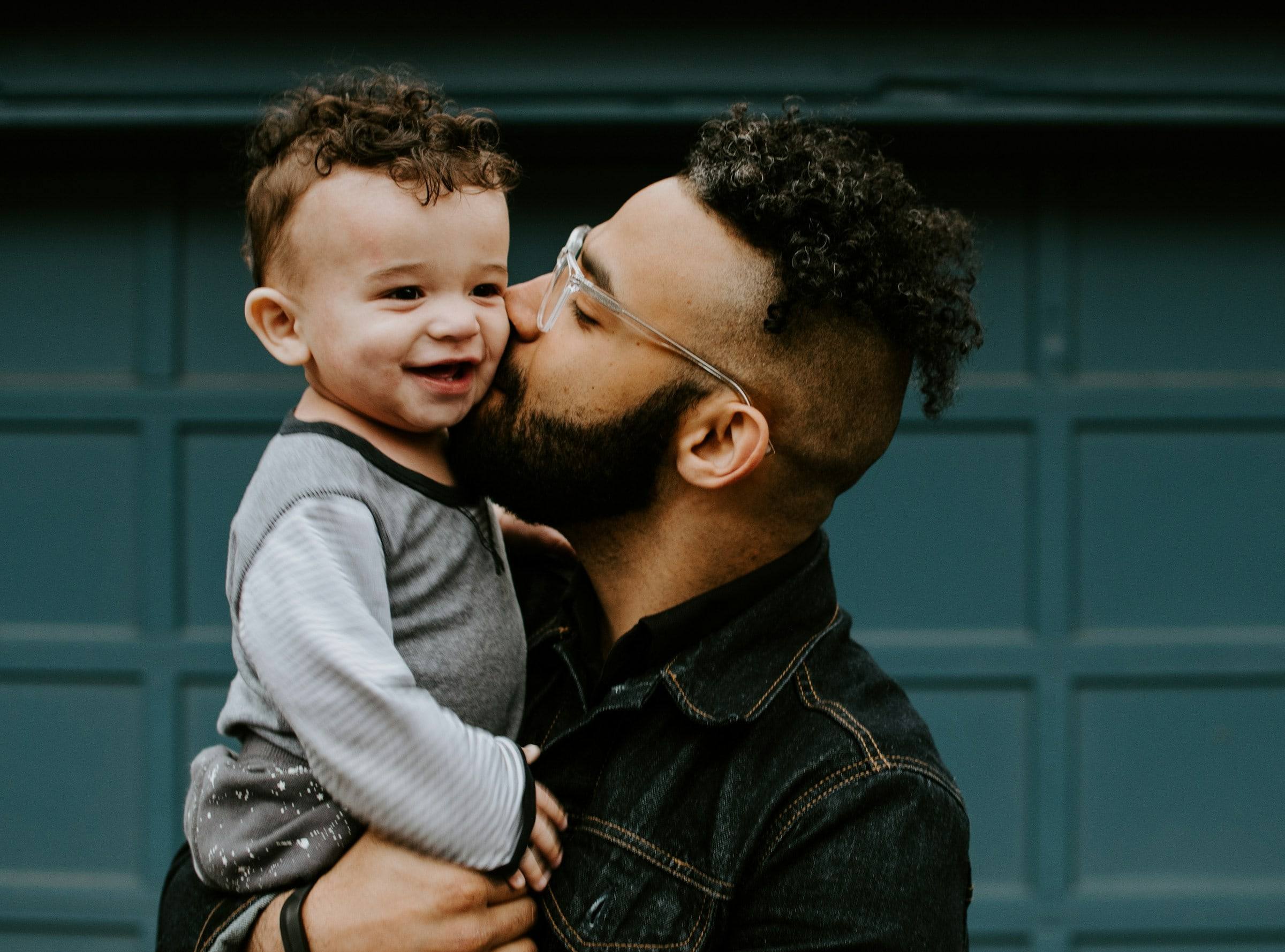 A father kissing the cheek of the laughing little boy in his arms.