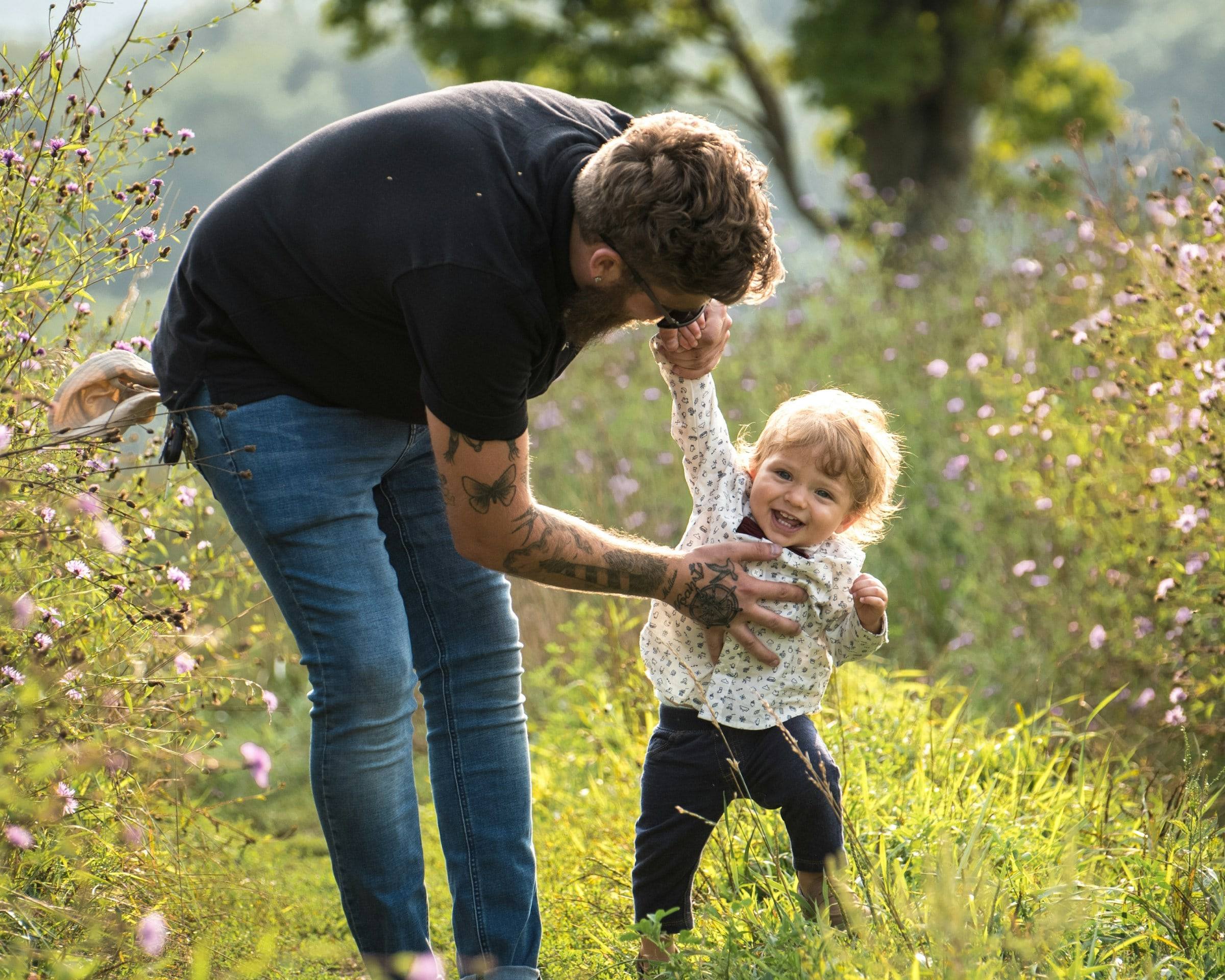 A father holding his son’s hands as he tries to walk across the grass.