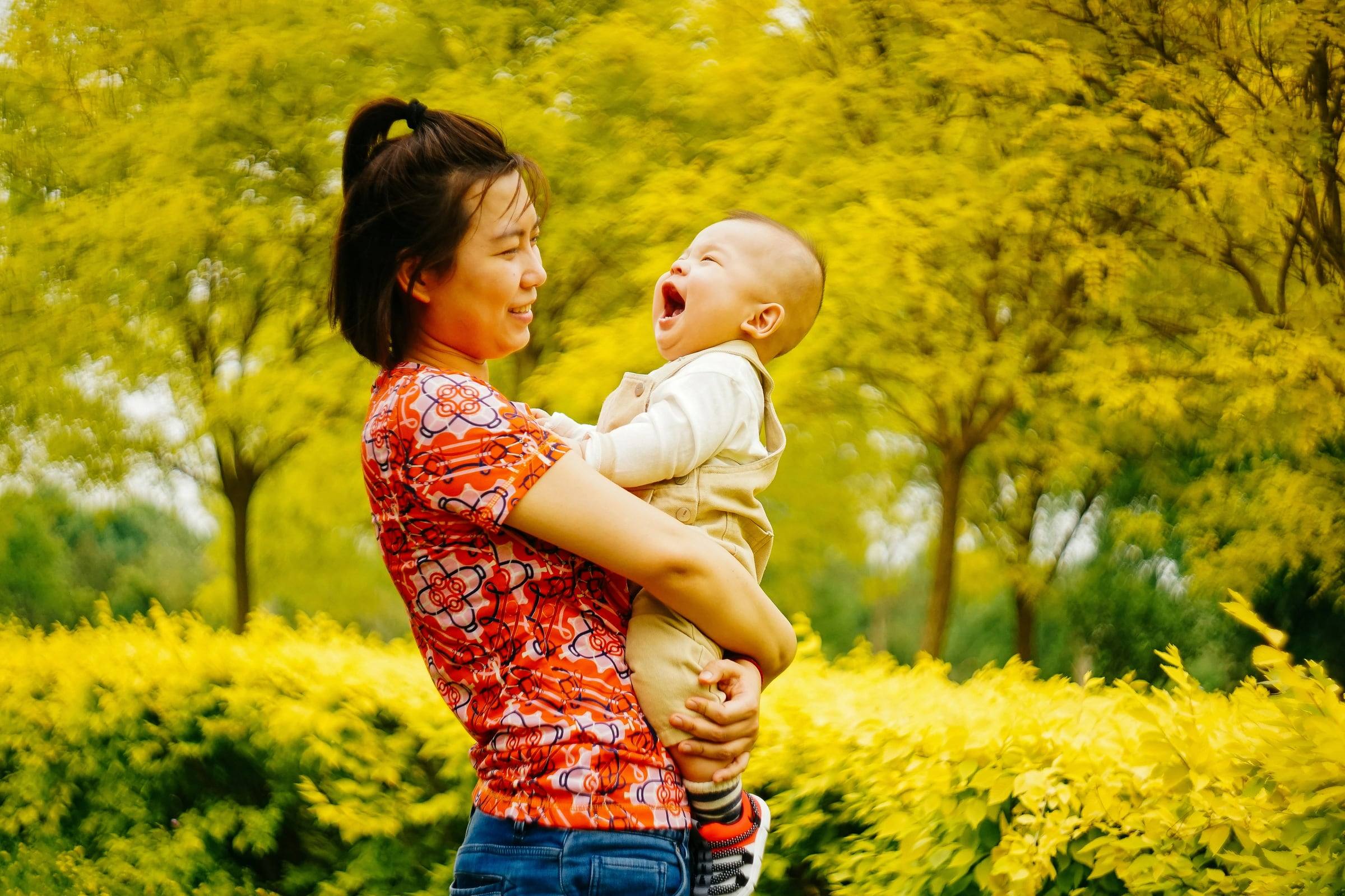 A mother and her baby laughing face to face in front of yellow-leaved trees.