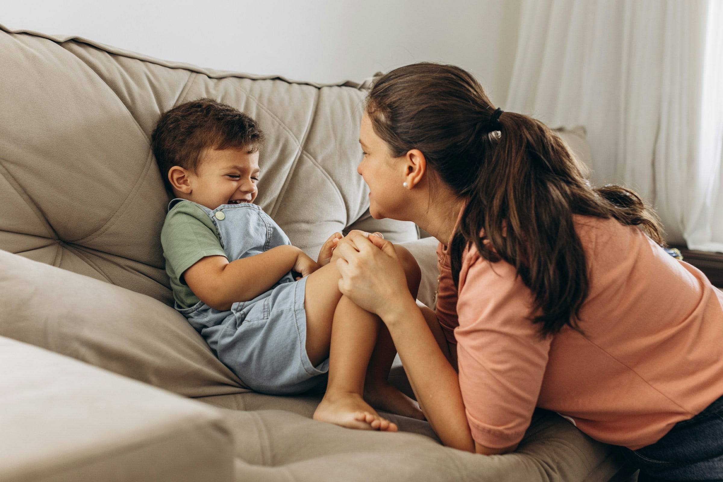 A little boy laughing on the sofa while his mother leans in close.