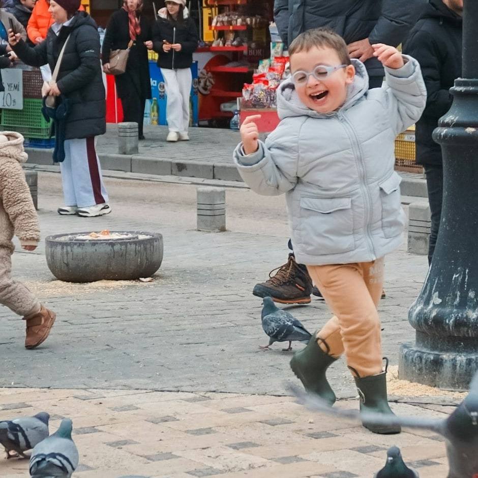A small boy in glasses running and laughing after pigeons in a square.
